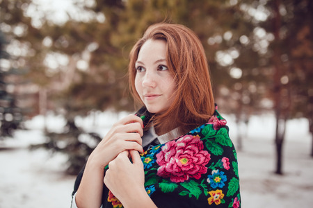 Russian beauty woman with red hair and freckles in the national scarf stands on a background of trees.の写真素材