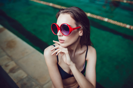 Beautiful brunette woman in funny sunglasses with a professional make up sitting near the pool.の写真素材