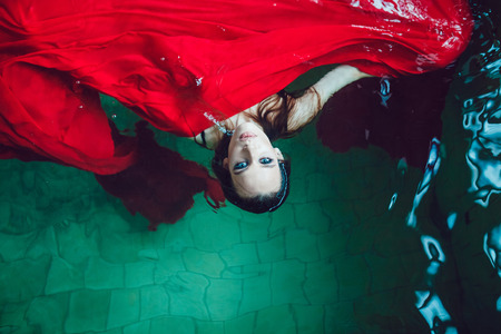 Fashion brunette woman with beautiful professional make up floating in a pool of red cloth.の写真素材