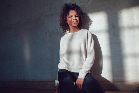 Beautiful African girl dressed in blank sweatshirt sitting on a gray background. Mock-up.の写真素材