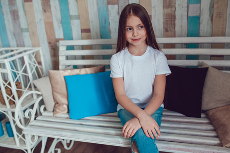 Beautiful little girl in blank white T-shirt and jeans is sitting on a wooden couch.の写真素材