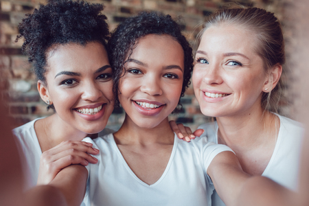 Beautiful and funny girls in white T-shirts makes selfie. Two afro-americans and one european.の写真素材