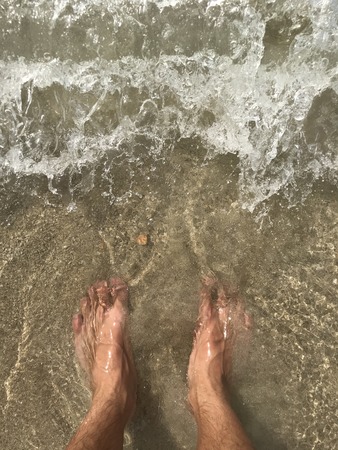 Top view of a mans leg standing on the sand and lapped by the ocean waves.の写真素材