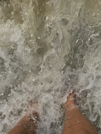 Top view of a mans leg standing on the sand and lapped by the ocean waves.