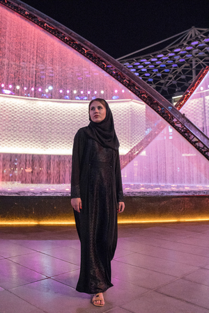 Muslim woman in a hijab stands against a background of fountains at night.