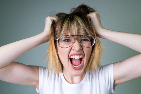 Emotional nervous girl with blond hair in transparent glasses, holding hands on head, making...