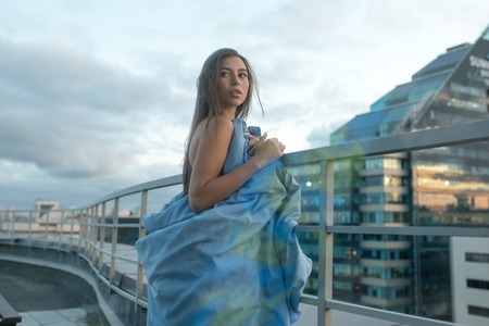Naked girl wrapped in a blanket stands on the terrace overlooking the city.の写真素材