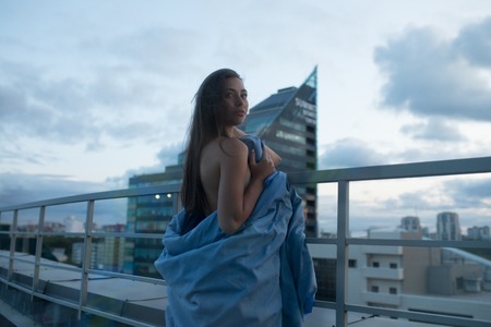 Naked girl wrapped in a blanket stands on the terrace overlooking the city.の写真素材