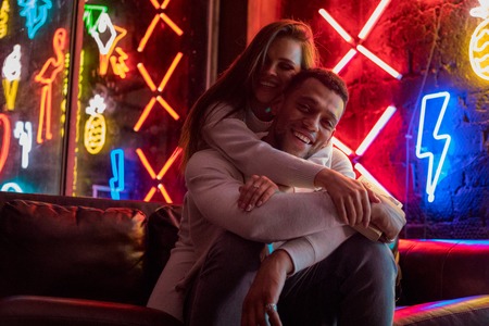 Happy multiracial couple sits embracing on the background of neon lamps.の写真素材