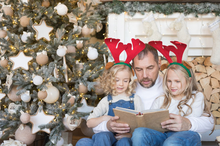 Dad reads daughters a book near the Christmas tree.の写真素材