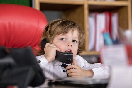 Little boy sits in the office in the chair and talking on the phone. Little boss.の写真素材