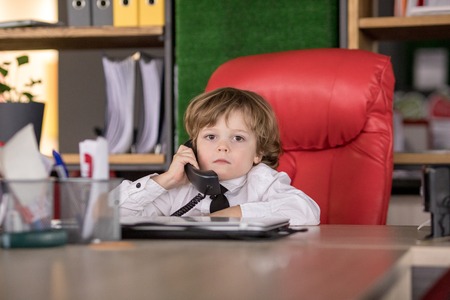 Little boy sits in the office in the chair and talking on the phone. Little boss.の写真素材
