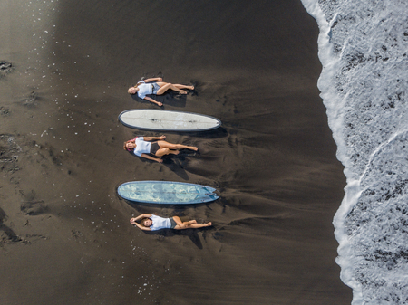 Top view of a women surfers with a board lying on the beach with black sand.