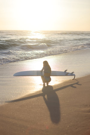 A Surfer Woman Can Be Seen Strolling Along the Beach as the Sun Sets in the Beautiful Skyの写真素材