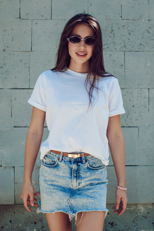 Young Woman In A White T-Shirt And Denim Skirt Posing Against Concrete Wallの写真素材