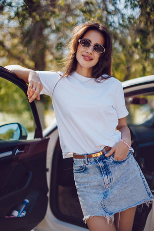 Young woman in white t-shirt and denim skirt standing by carの写真素材