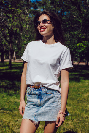 Smiling Young Woman In A White T-Shirt And Denim Skirt Standing Outdoors In Parkの写真素材