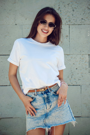 Young Woman In A White T-Shirt And Denim Skirt Posing Against Concrete Wallの写真素材