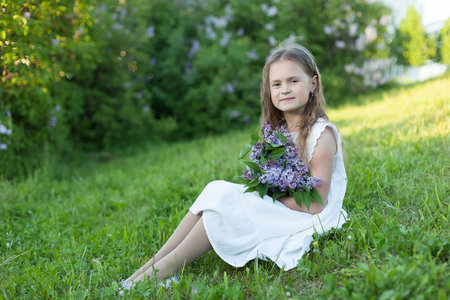 A little girl holds a bouquet of lilacs on a beautiful Sunny day. Cute girl outdoors in a blooming gardenの写真素材