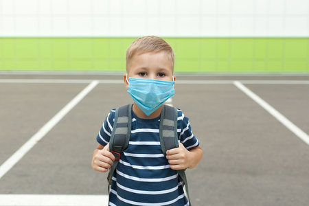 Little blond schoolboy wearing mask during corona virus outbreak. Little kid wears blue backpack, breathes through mask, going to school. Back to school concept after reopening. Child near school wallの写真素材