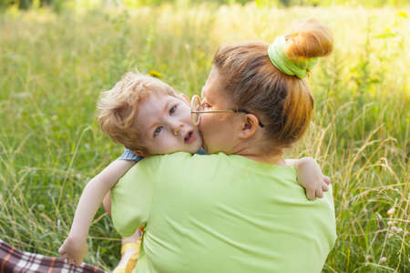 A happy mother and a disabled child are sitting on the grass in the park and gently hugging. Summer holidays. Disability.の写真素材