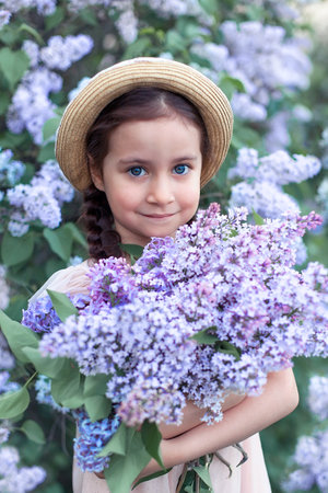a beautiful little girl in a straw hat on her head stands in the lilac bushes and holds a bouquet in her hands, looks towards the camera and smiles.A brooding and May evening sunsetの写真素材