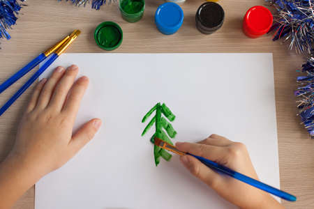 Childrens hands of a girl dip a brush in paint. In the background there are brushes, paints and New Years tinsel. Paints on the table. A child draws and makes crafts in kindergarten.の写真素材