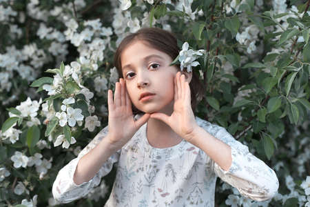 A sweet 9-year-old girl with big beautiful eyes and a light dress in blooming white apple trees....