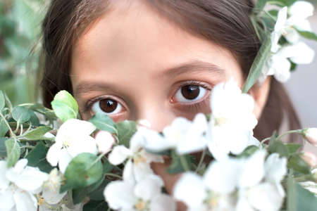 A little beautiful teenage girl in a white dress and sweater enjoys the spring flowering of apple trees, smiles and looks at the camera. Spring.の写真素材