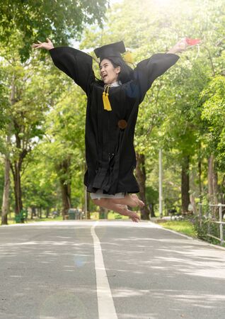Happiness Young asian woman student in her graduation gown and wearing graduation cap with certificate in hands.Young female happy and jumping with Graduate day.Education finish congratulation conceptの写真素材