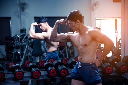 Asian handsome young muscular asian man exercising bodybuilder  in front of a glass in the gym .Guy flexing trains his bicep muscles, showing body at the camera in the gym.Exercise in gym concept.の写真素材