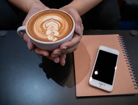Hand woman holding a cup of hot latte arts coffee with cell phone and note on black table. Barista coffee art concept.with copy spaceの写真素材