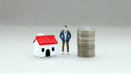 A miniature man standing between a pile of coins and a miniature house.の写真素材