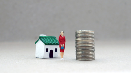 A miniature woman standing between a pile of coins and a miniature house.の写真素材