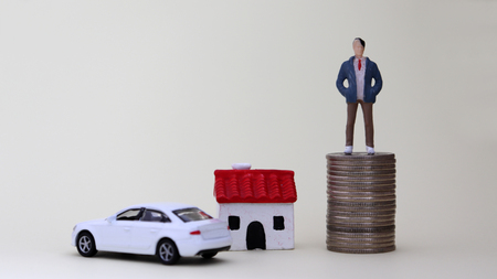 A miniature man standing on a pile of coins. A miniature house and a miniature car.の写真素材