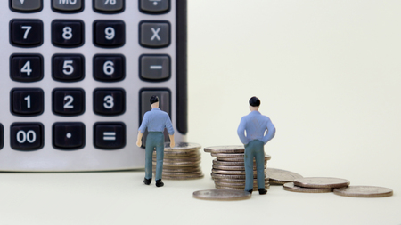 Miniature men standing next to a pile of coins and a calculator.の写真素材