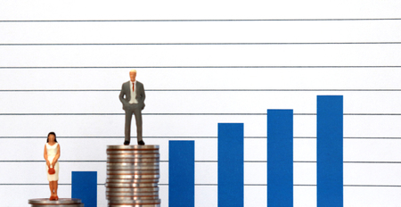 A miniature man and a miniature woman standing on top of a pile of coins at different heights in front of a bar graph.の写真素材