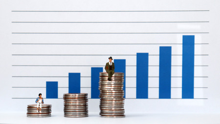 A miniature man and a miniature woman sitting on a pile of coins at different heights against a bar graph.の写真素材