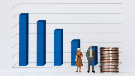 A miniature man and woman in a suit with a pile of coins standing in front of a three dimensional bar graph.の写真素材