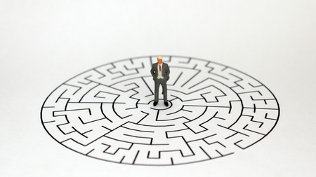 A miniature man standing in the middle of a maze on a white background. The male concept in agony.