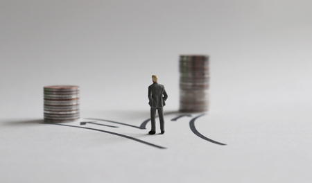 A miniature man standing in front of two paths with a stack of coins of different heights.の写真素材