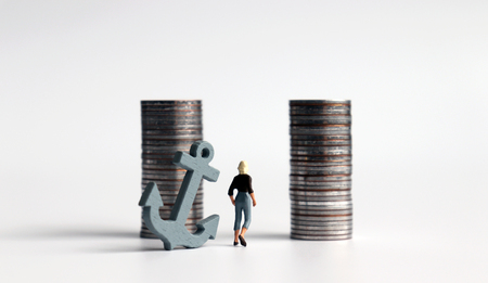 A miniature woman with an anchor trying to walk between two piles of coins.の写真素材