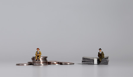 A miniature man sitting on a pile of coins and a miniature man sitting on a pile of bills.の写真素材