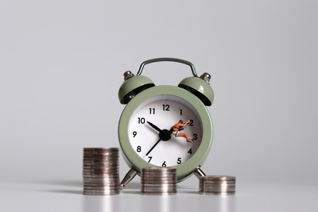 A miniature person with an alarm clock and a pile of coins. A concept of working hours and income...