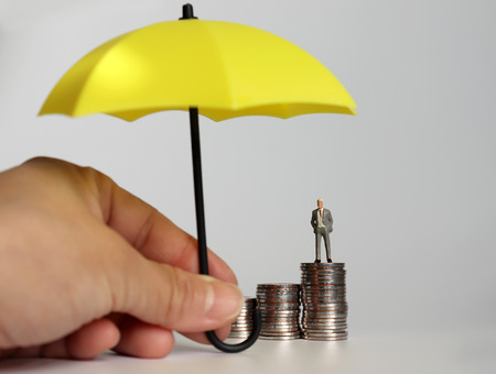 A hand holding a yellow umbrella and a miniature man standing on a pile of coins.