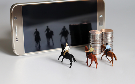 Miniature man riding a horse. Smartphone and pile of coins.の写真素材