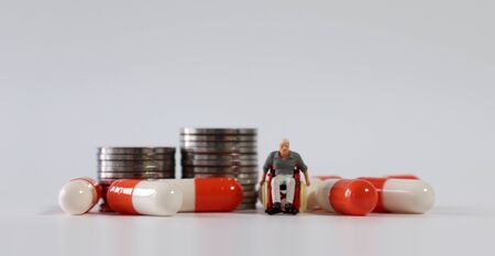 A miniature man in a wheelchair and pills around him in front of a pile of coins.の写真素材