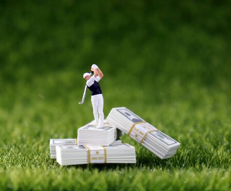 A miniature golfer standing on a wad of hundred dollar bills with a lawn background.の写真素材