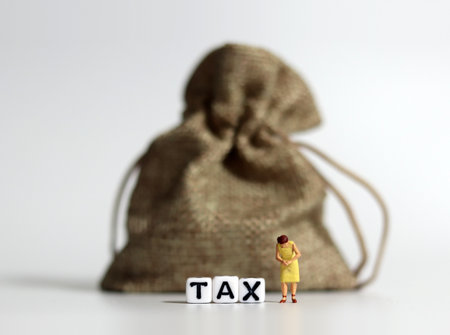 White cube bearing the word TAX with a bundle and a miniature woman standing with her head down. Concepts about the difficulty of paying for rising taxes.の写真素材