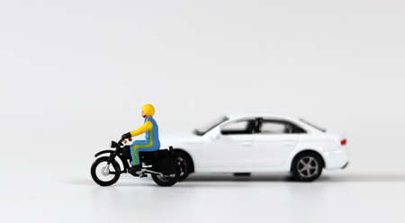 A miniature driver wearing a helmet next to a white miniature car and riding a motorcycle. Miniature...